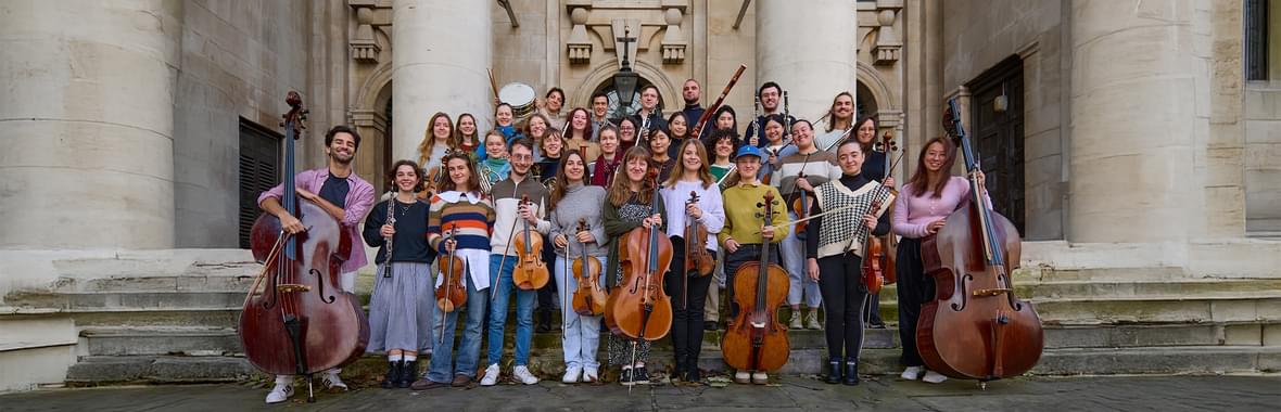 Orchestra standing together holding their instruments in front of a white stone historical building.