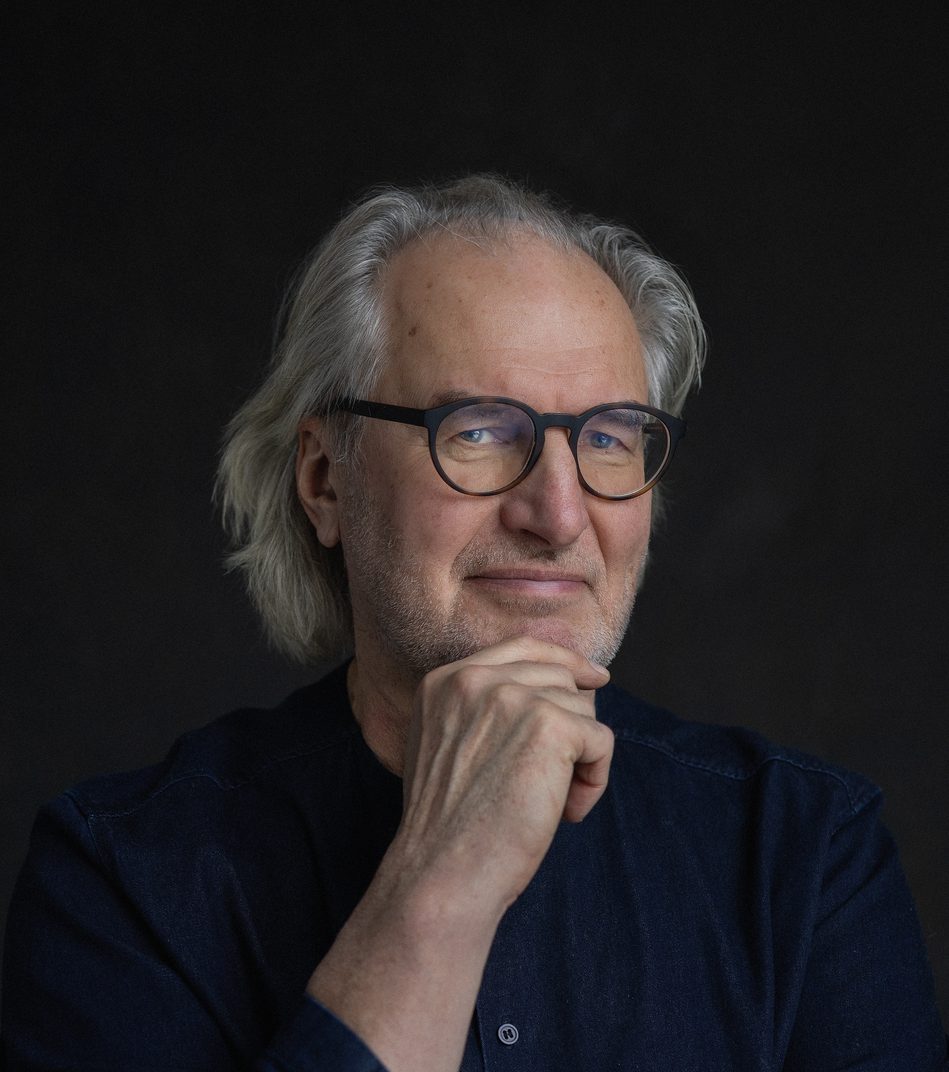 Man with white hair wearing glasses against a dark background looks at the camera with an enigmatic smile.