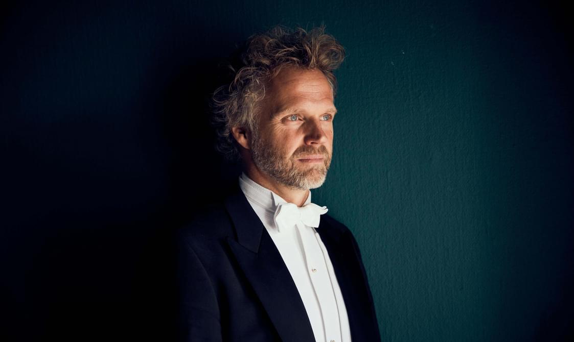 Man with short curly hair and a beard wearing a black suit and white bow tie looking away from the camera.