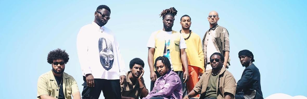 Group of men sitting and standing in a decorated boat against a bright blue sky.