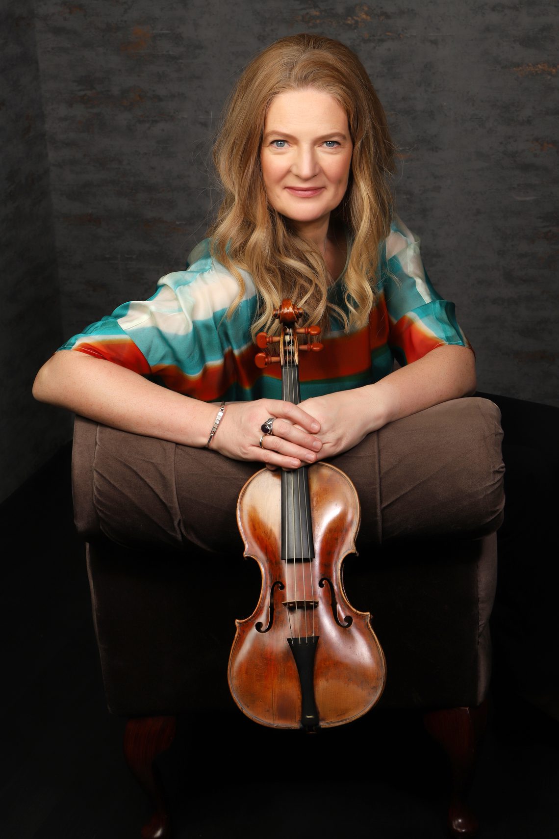 Woman with long blonde hair sitting and facing backwards to the camera, holding a violin.