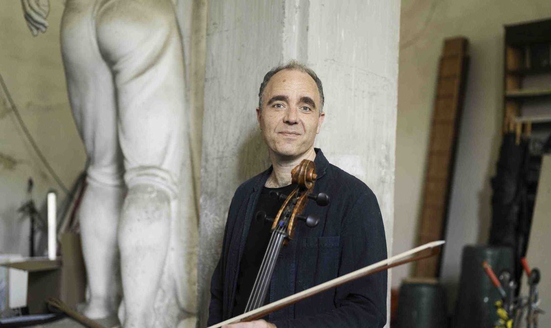 Man with cropped hair holding a cello and standing in front of an ancient stone pillar.
