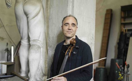Man with cropped hair holding a cello and standing in front of an ancient stone pillar.