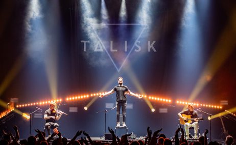 Man standing on a stage with lights and smoke effects, flanked by a man playing a violin and another man playing guitar.