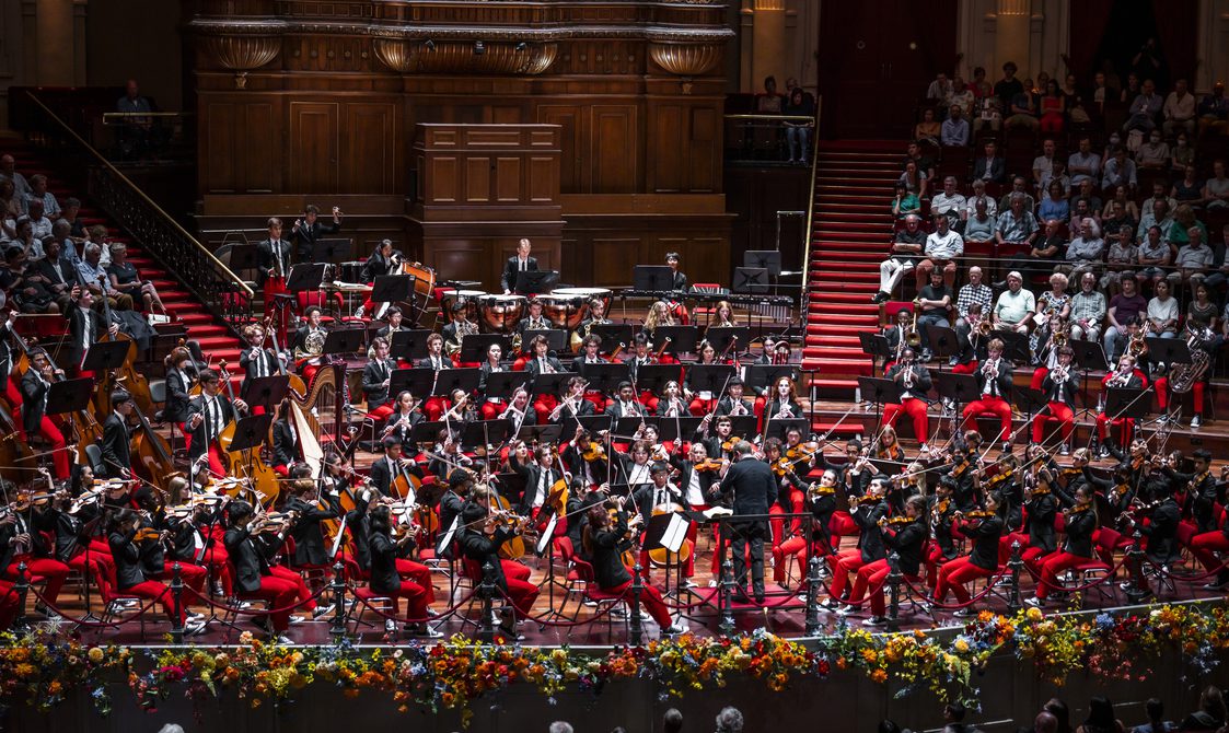 Full orchestra onstage wearing dark jackets and red trousers.
