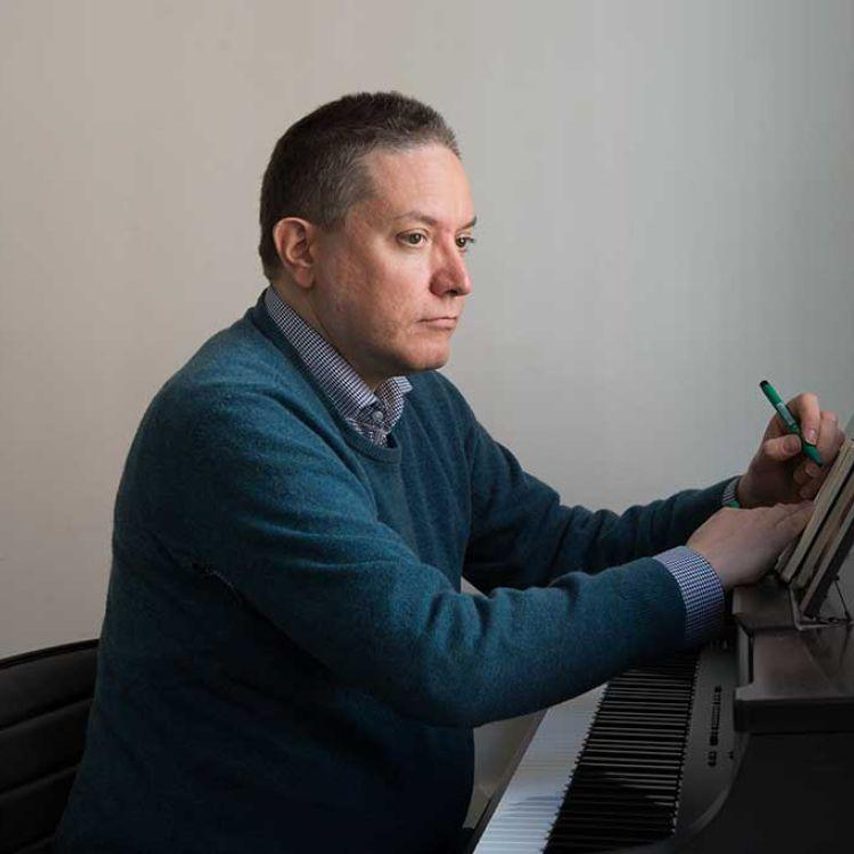 Man with short grey hair sitting at a piano looking pensive.