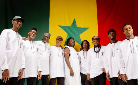 Group of eight men and one woman wearing white tunics and caps, standing in front of a Senegal flag.