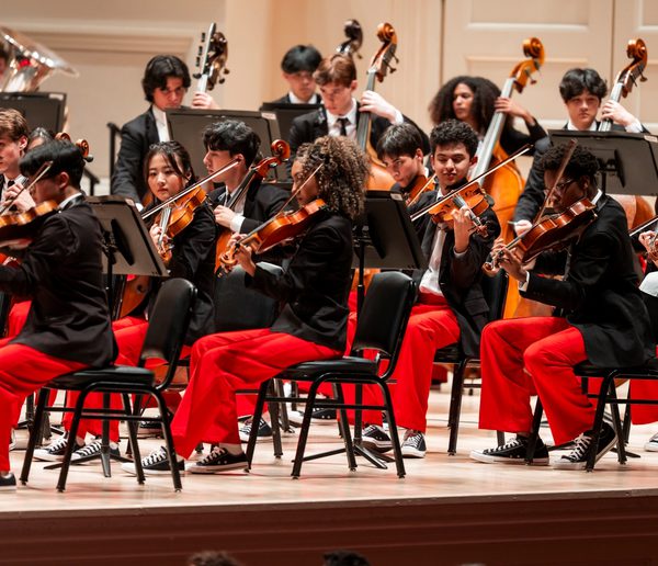 Group of musicians onstage playing instruments wearing black jackets, red trousers and trainers.