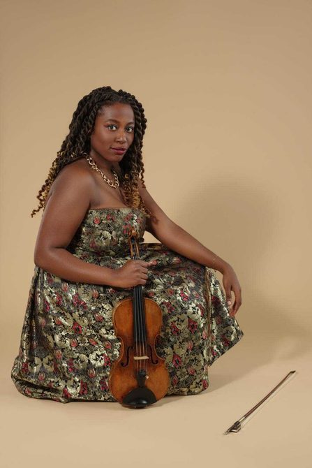 Black woman with long dark hair wearing a floral dress and holding a violin.