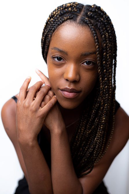 Close-up of Black woman with braided hair holding her hands beside her face and looking into the camera.