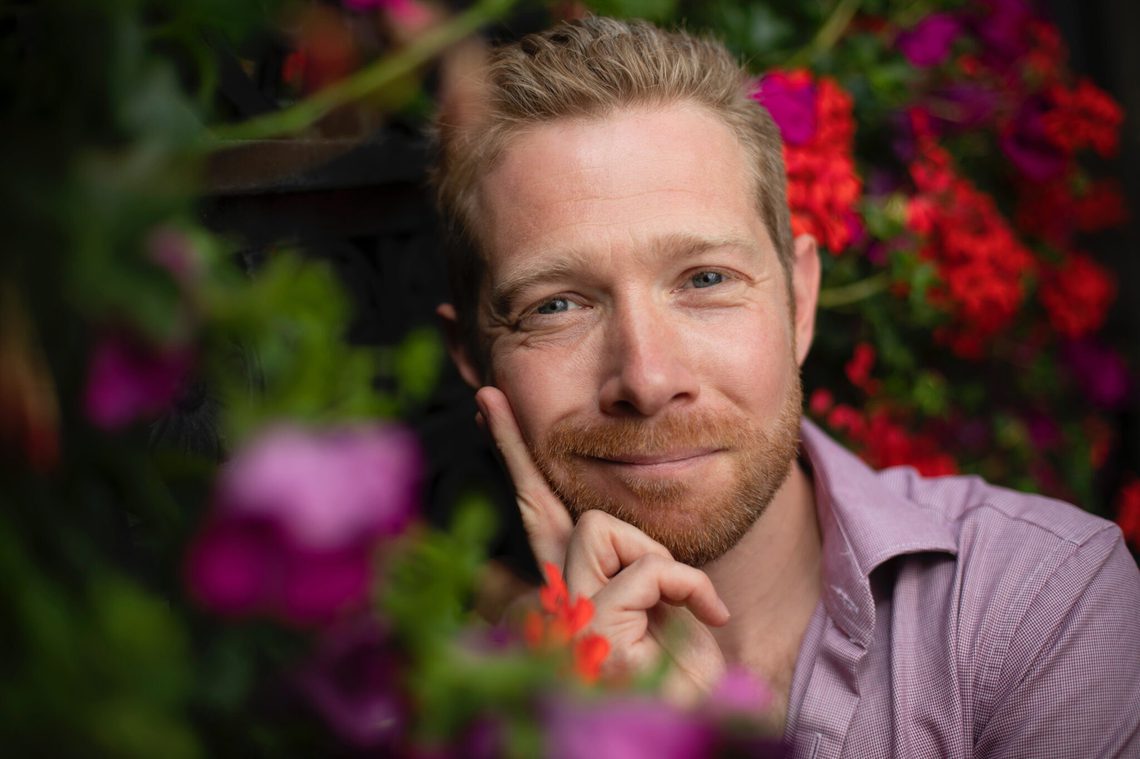 Close-up of man with blond hair and a beard, surrounded by flowers and looking into the camera.