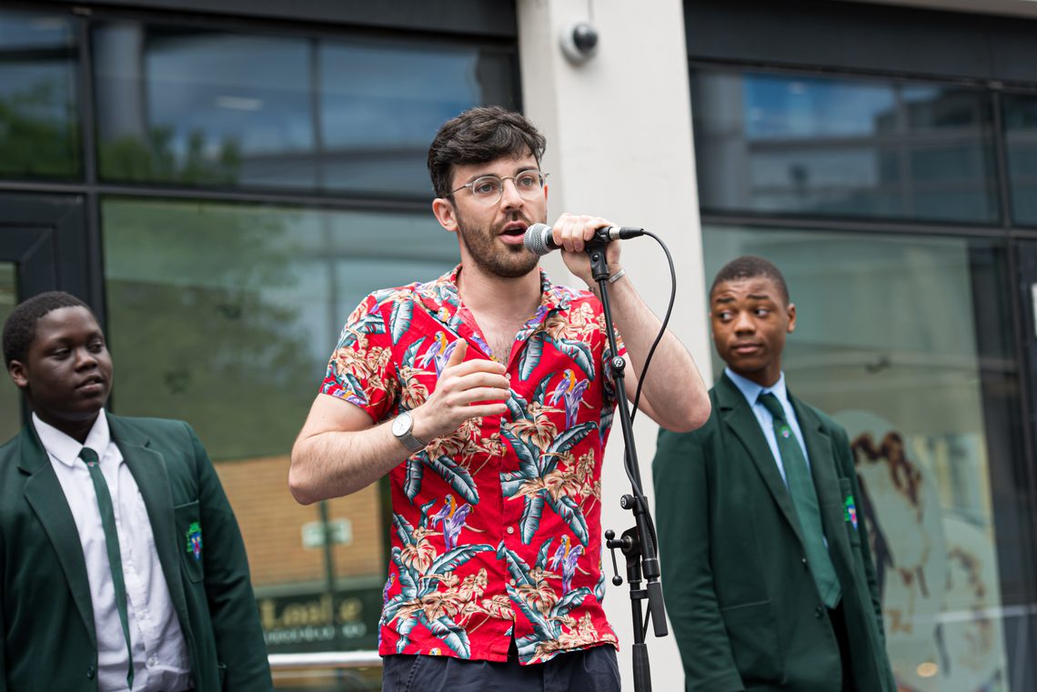 Man in Hawaiian shirt singing into a microphone with two boys in school uniform standing behind him