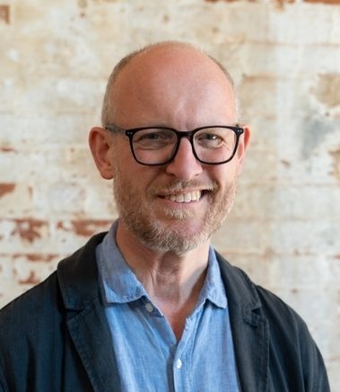 Man with shaved head and a beard wearing a blue jacket and glasses, smiling at the camera.