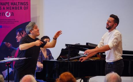 Two men standing beside a grand piano, one man is conducting, the other is singing