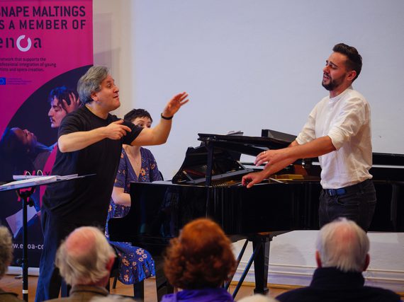 Two men standing beside a grand piano, one man is conducting, the other is singing