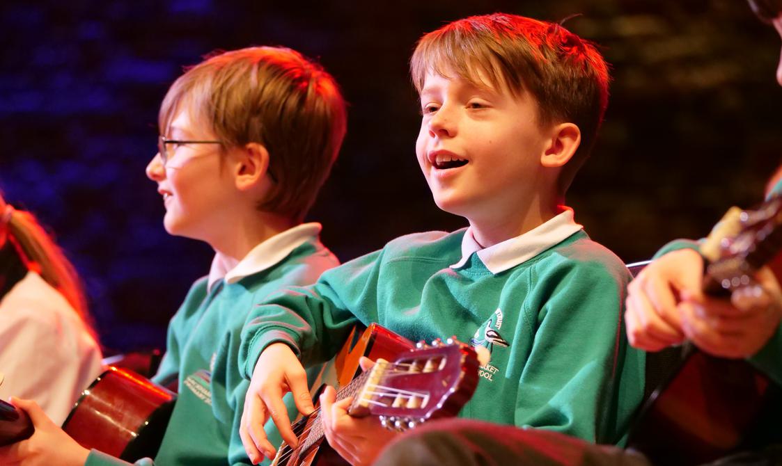 Small boy wearing a green sweatshirt singing and playing guitar.
