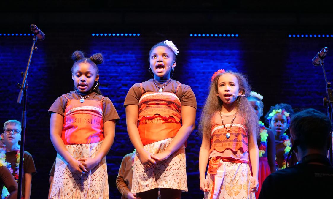 Three young girls wearing colourful costumes and singing onstage.