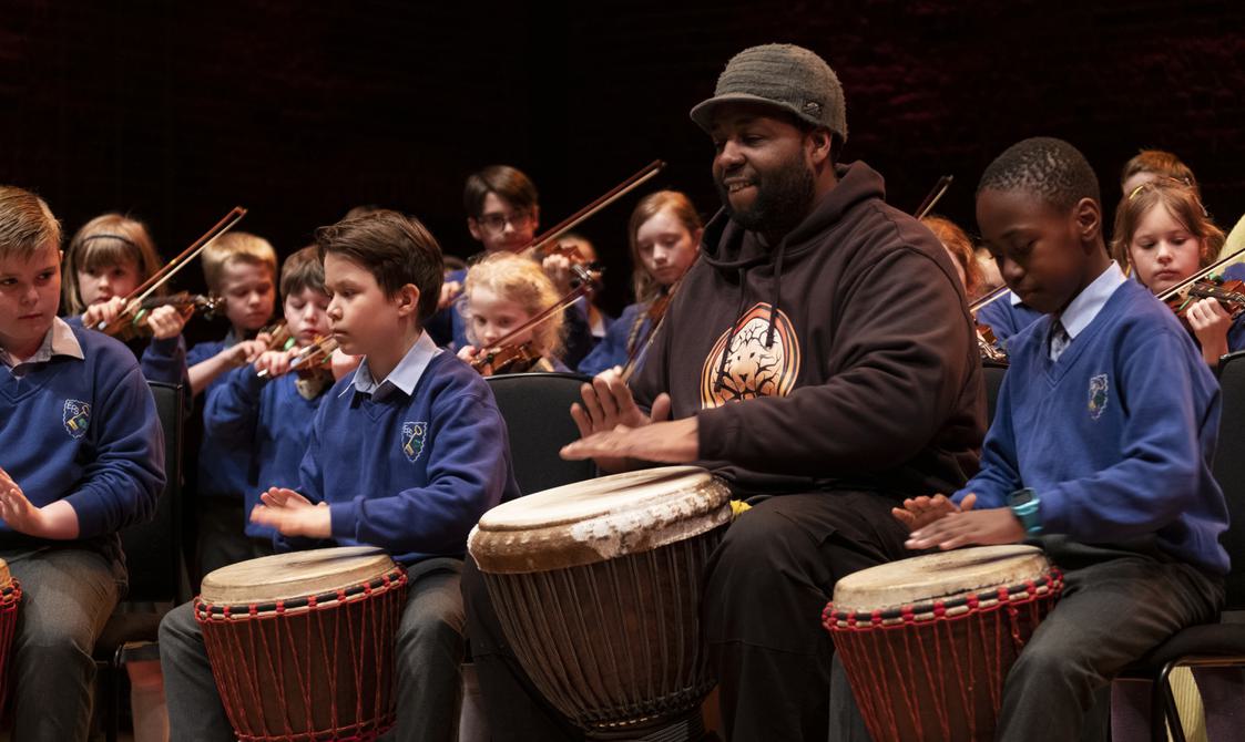 Man wearing a cap and hoodie playing drums with a group of schoolchildren.