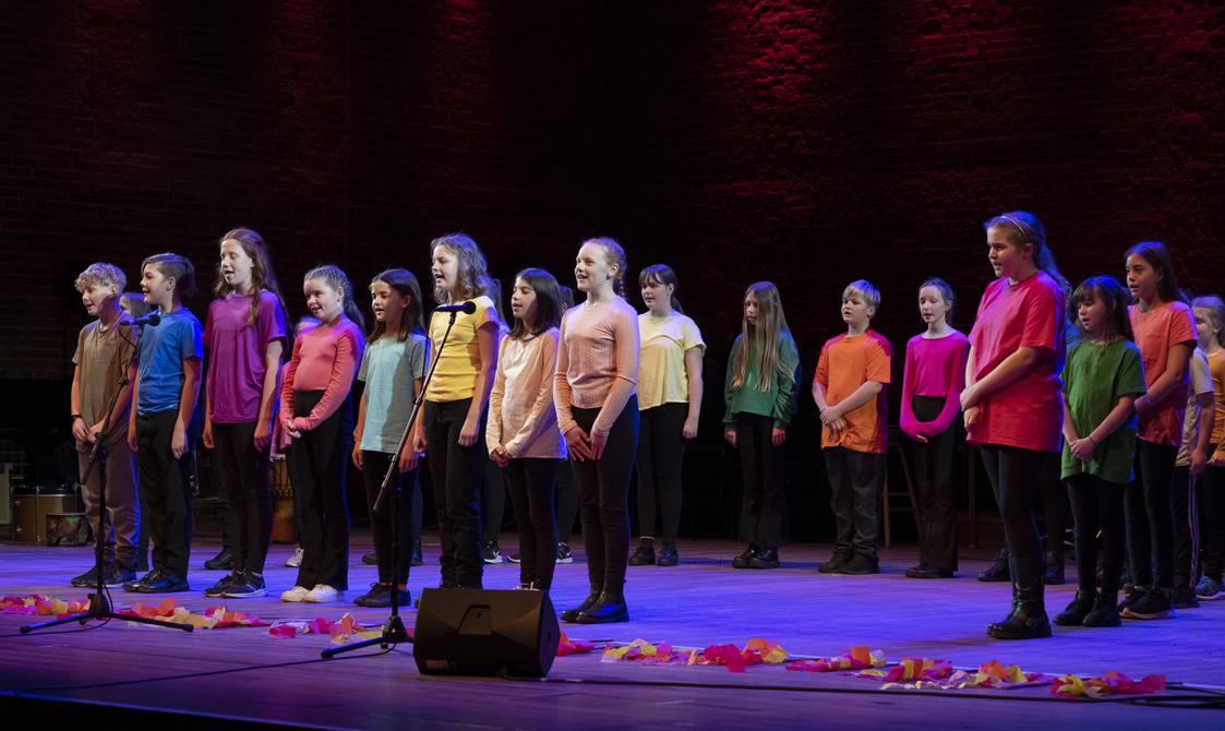 Stage full of children wearing t-shirts in a range of colours and singing.