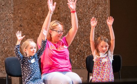 Woman in a pink t-shirt sitting on a chair, between two young girls, all of them holding their arms up in the air.