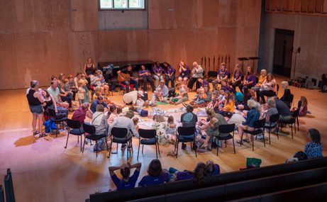 Large group of adults and children sitting in a circle in a hall with musical instruments
