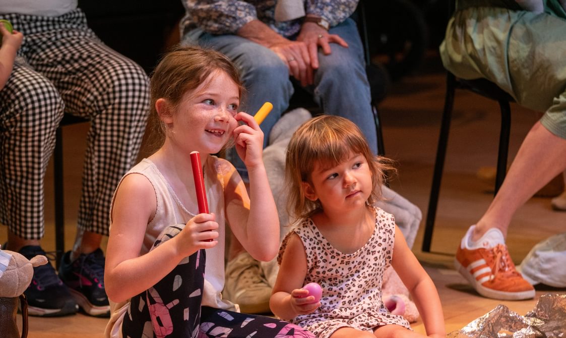 Two young girls sat on a wooden floor. The girl on the left is holding two brightly coloured wooden sticks and smiling, and is wering a grey tank top, leggings with flowers on, and pink sandals. The girl on the right is younger, and she is wearing a leopard print dress, pink sandals and holding a pink ball. In the background you can see the legs of adults sat on chairs behind them.