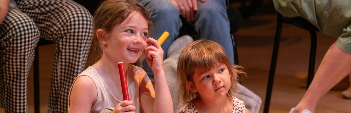 Two young girls sat on a wooden floor. The girl on the left is holding two brightly coloured wooden sticks and smiling, and is wering a grey tank top, leggings with flowers on, and pink sandals. The girl on the right is younger, and she is wearing a leopard print dress, pink sandals and holding a pink ball. In the background you can see the legs of adults sat on chairs behind them.
