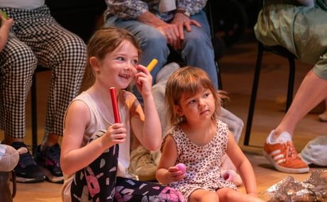 Two young girls sat on a wooden floor. The girl on the left is holding two brightly coloured wooden sticks and smiling, and is wering a grey tank top, leggings with flowers on, and pink sandals. The girl on the right is younger, and she is wearing a leopard print dress, pink sandals and holding a pink ball. In the background you can see the legs of adults sat on chairs behind them.