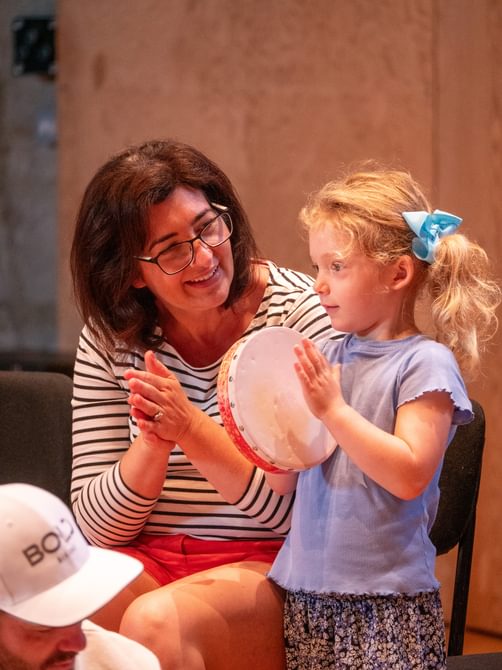 Woman smiles and claps while watching a young girl playing the tambourine