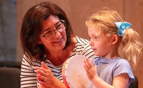 Woman smiles and claps while watching a young girl playing the tambourine