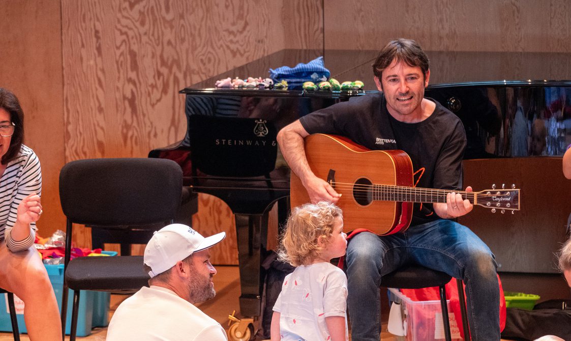 Man sits in front of a piano playing the guitar and singing while a group of adults and children play around him