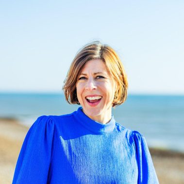 Woman with a blond bob wearing a bright blue top, standing on the beach and smiling.