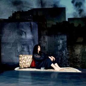 Woman sitting on stage, leaning against a patterned cushion.