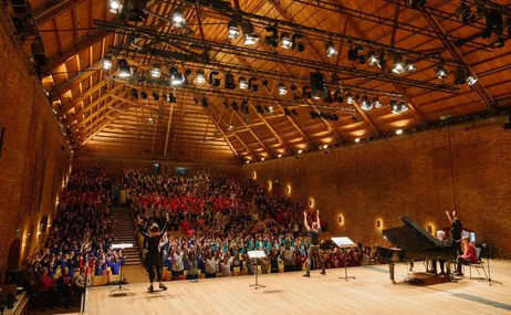 A group of children in a hall. There is a stage with some musicians on.