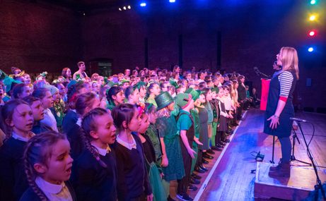 A person on stage singing to an audience of children who are also singing.