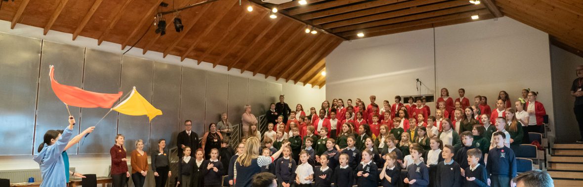 A person on a piano and a group of children singing in a hall.