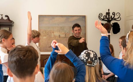 A teacher pointing towards a group of schoolchildren with their hands up.