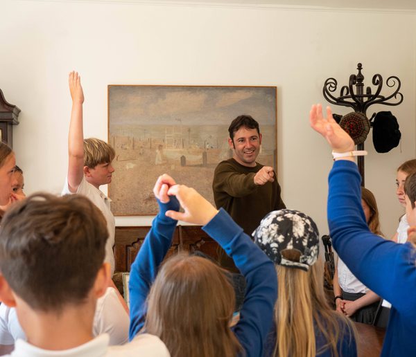 A teacher pointing towards a group of schoolchildren with their hands up.