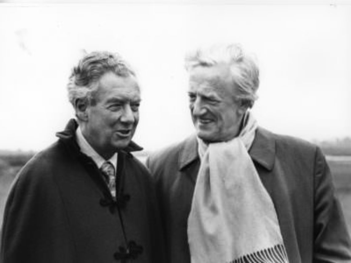 Two middle aged men in outdoor coats and light scarves, walking and laughing together