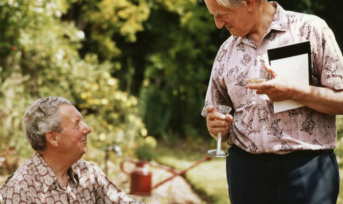 One man seated, the other standing, looking lovingly at each other. They are outside, in summer shirts.