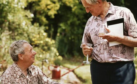 One man seated, the other standing, looking lovingly at each other. They are outside, in summer shirts.