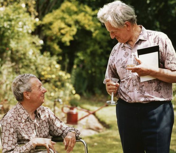 One man seated, the other standing, looking lovingly at each other. They are outside, in summer shirts.