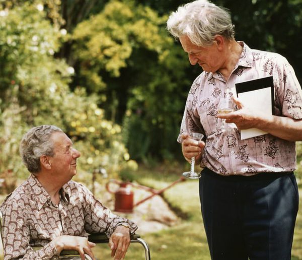 One man seated, the other standing, looking lovingly at each other. They are outside, in summer shirts.
