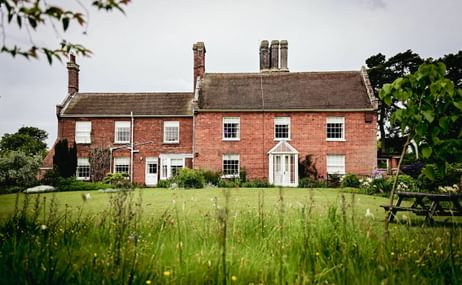 A red house with white windows, a big lawn in front of it and long grass in front of the camera. There is a bench on the right hand side, and some leaves coming down in the top left corner.