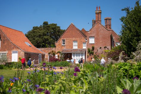 Red brick farmhouse with people strolling outside, greenery and flowers in the foreground.