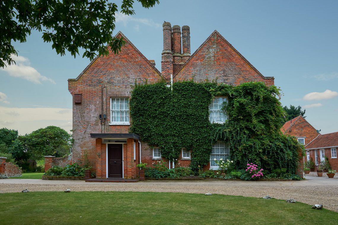 Side view of large red brick building covered in green climbing plants, under a blue sky.