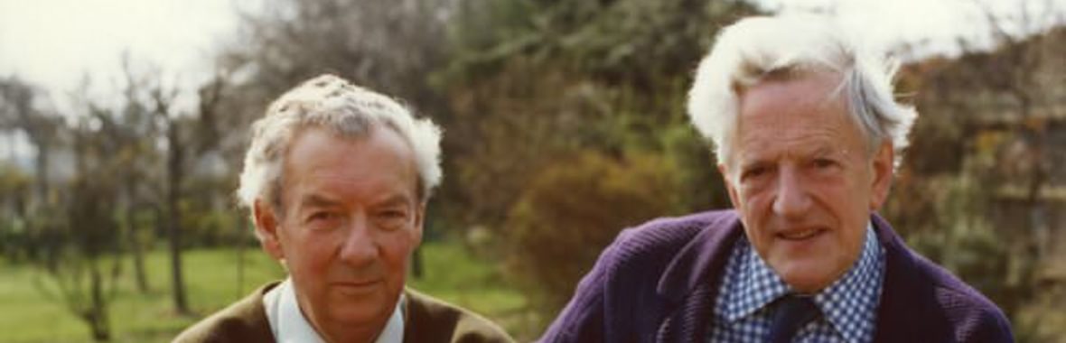Portrait of two older men with grey hair, in cardigan suits, on outdoor chairs, sitting close together