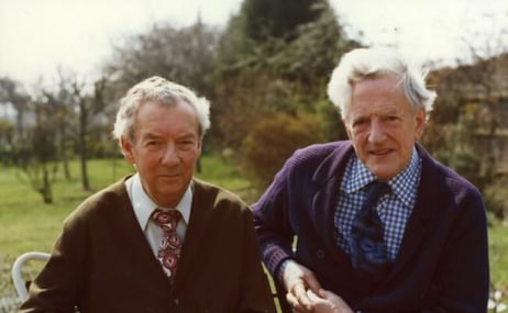 Portrait of two older men with grey hair, in cardigan suits, on outdoor chairs, sitting close together