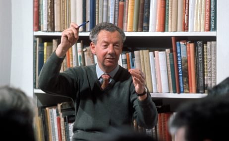 A man with grey curls 'conducts' with a pencil, with rows of books in the background