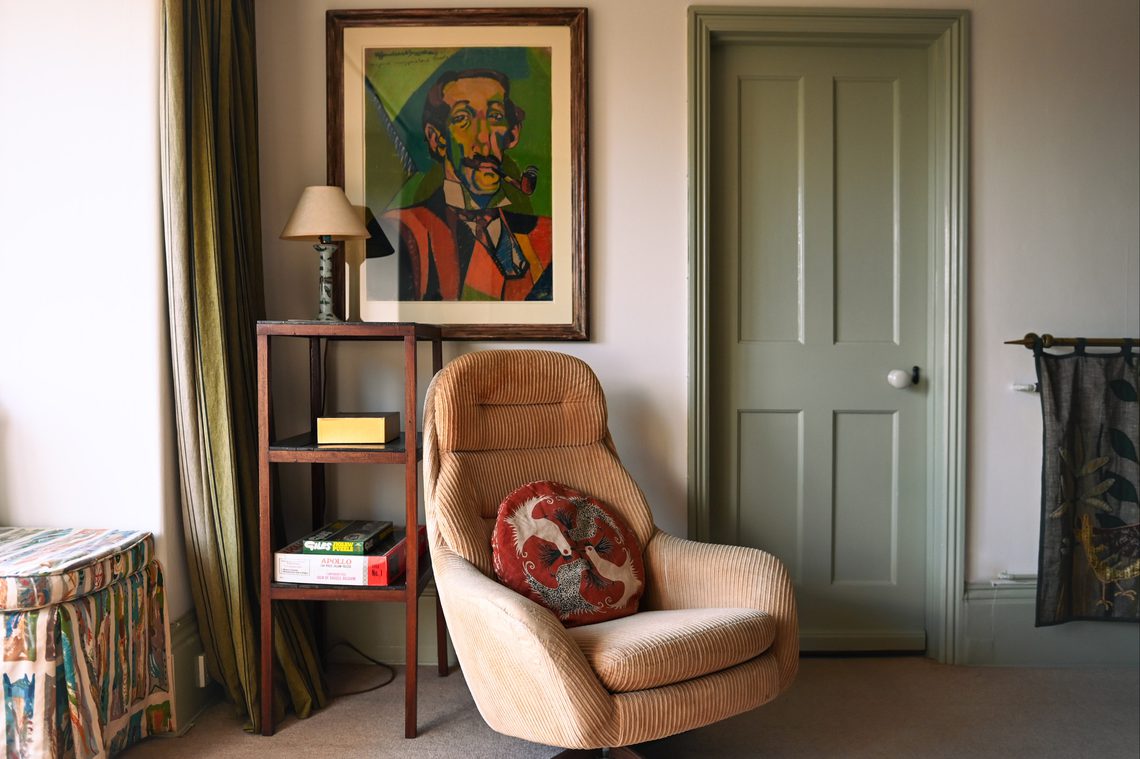 Living room scene with cosy armchair in the centre, bookshelf with table lamp, artwork and green door in the background